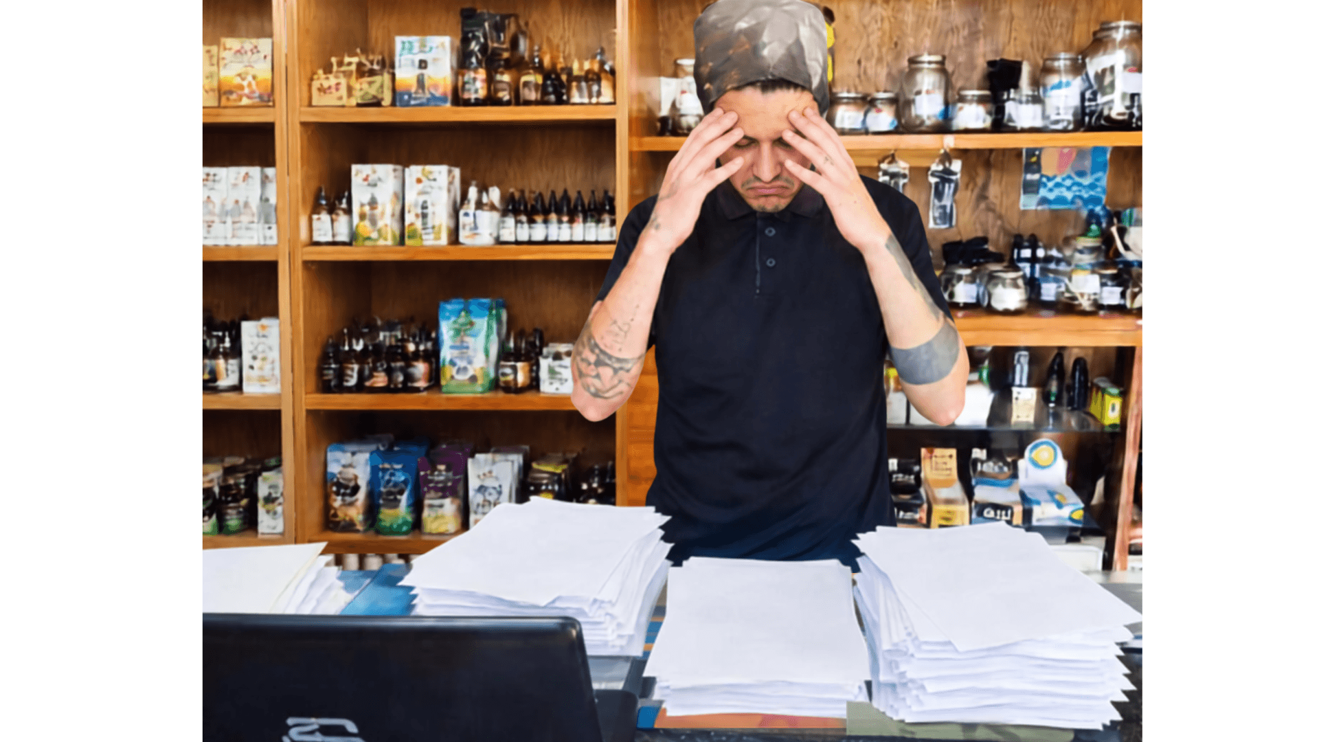 Stressed person surrounded by stacks of paperwork at desk in cannabis club with shelves of products behind them.