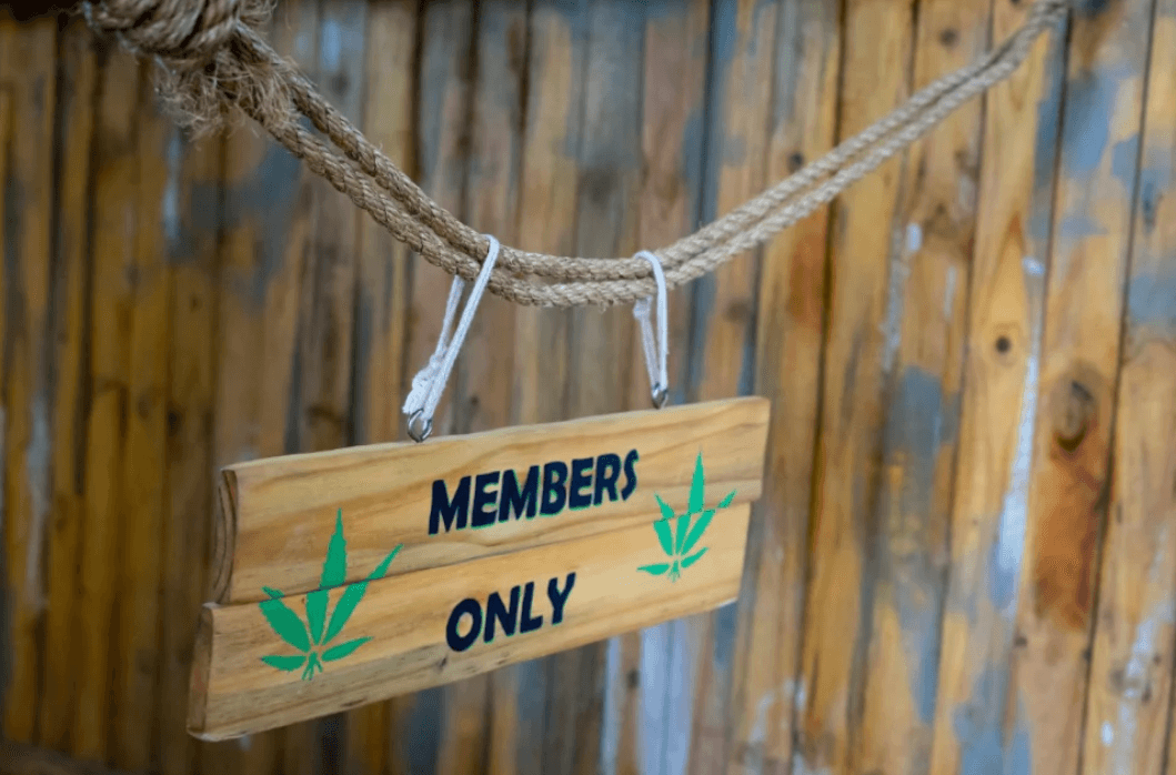 Wooden "Members Only" sign with cannabis leaf graphics hanging from rope against wooden fence.
